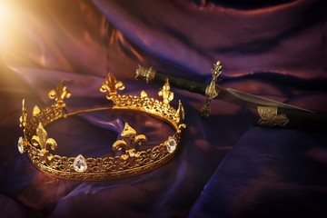 low key image of beautiful queen/king crown and sword over dark royal purple delicate silk. fantasy...