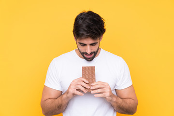Young handsome man with beard over isolated yellow background eating a chocolate tablet
