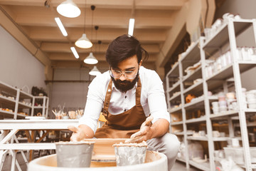 A young male potter is engaged in craft in his workshop on a potter's wheel and makes a clay product