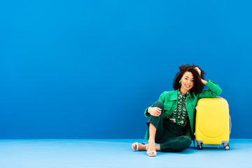 smiling african american woman sitting near travel bag on blue background