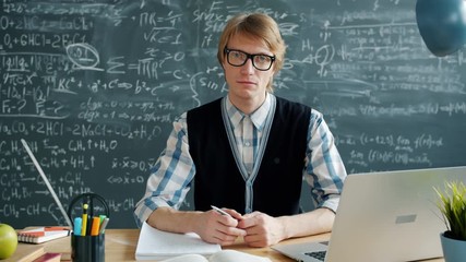Portrait of serious handsome young man in glasses looking ay camera sitting at desk in classroom alone. Modern education, people and occupation concept.