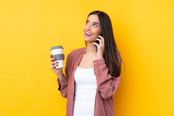Young brunette woman over isolated yellow background holding coffee to take away and a mobile