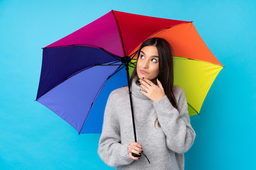 Young brunette woman holding an umbrella over isolated blue wall thinking an idea