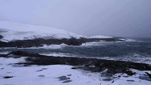 Barents Sea. Waves hit a rocky snowy shore. Snow flies into the camera. Overcast weather. The smooth movement of the camera.