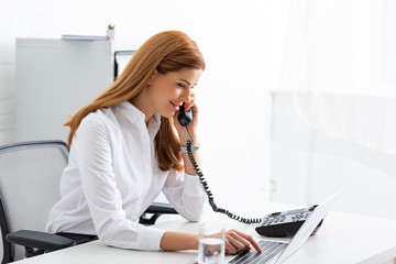 Side view of smiling businesswoman talking on telephone and using laptop on table