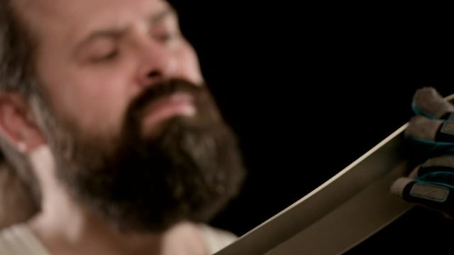 Man with beard, gloves in black studio, runs his fingers along blade of machete.