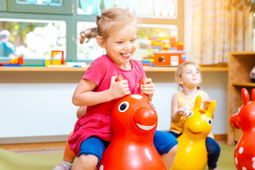 Little girls riding on play horses in kindergarten