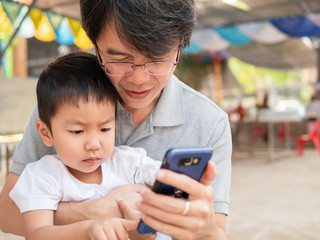 Fototapeta premium Asian little child boy watching smart phone with dad with happy face. Kid and father looking to mobile phone together outdoor.