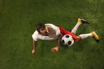 Top view of caucasian football or soccer player on green background of grass. Young male sportive model training, practicing. Kicking ball, attacking, catching. Concept of sport, competition, winning.