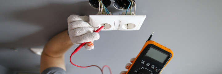 Man in hard hat and protective gloves checking wal outlet with tester