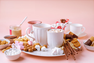 Hot chocolate in mugs, marshmallows, chocolates and cookies on pink background