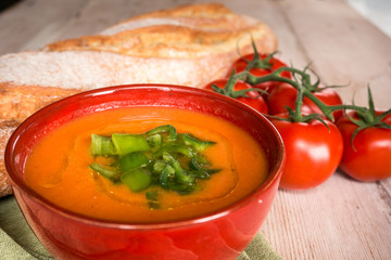 Cuenco rojo de gazpacho sobre mesa de madera clara, acompañado de tomates y pan