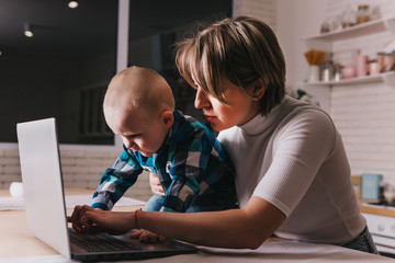Beautiful business mom is using a laptop while spending time with her cute baby boy at home