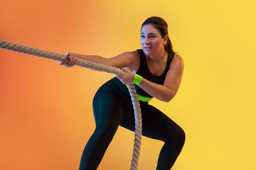 Young caucasian plus size female model's training on gradient orange background in neon light. Training upper body with the fit ropes. Concept of sport, healthy lifestyle, body positive, equality.