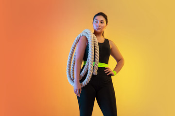Young caucasian plus size female model's training on gradient orange background in neon light. Posing confident with the fit ropes. Concept of sport, healthy lifestyle, body positive, equality. - Powered by Adobe