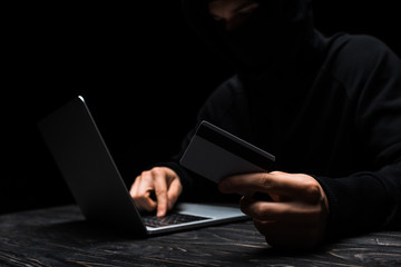 selective focus of hacker in mask using laptop while holding credit card isolated on black