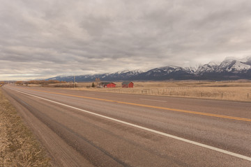 Red house and barn on side of empty highway with open pasture and snow capped mountain range