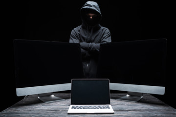 hacker in mask standing with crossed arms near computer monitors and laptop with blank screen ...