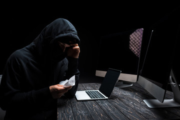 upset hacker holding tissue near laptop and computer monitors with blank screen isolated on black
