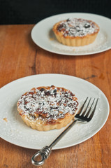 Top view, close distance of a freshly baked, cranberry raisin tart on with sprinckled ,powder sugar, on a round, white plate with an artisan fork on a wood serving board