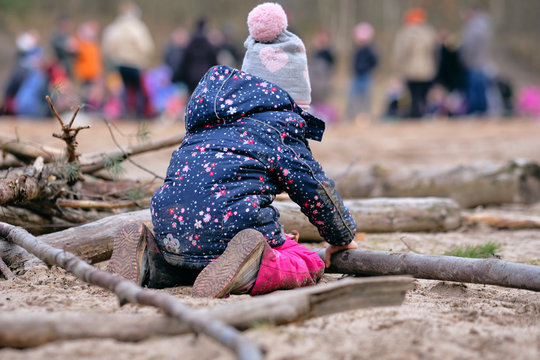 Rear View Of A 4 Year Old Child Girl In Warm Clothing Kneeing On The Ground  And Building Something With Wooden Branches In Front Of A Group Of People. Seen In Germany In February
