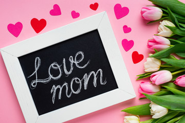 Top view of chalkboard with love mom lettering near paper hearts and tulips on pink background