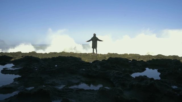 Man dressed in black raised his arms in front of the waves with the clear sky and a rainbow in slow motion