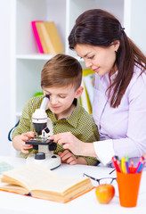 The boy and teacher are sitting at the table and looking at the microskim. Before the teacher lies...