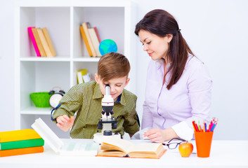 The boy and teacher are sitting at the table and looking at the microskim. Before the teacher lies...