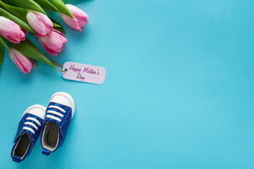 Top view of tulips with happy mothers day lettering on paper label near baby booties on blue background