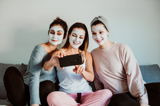 Group of friends applying a revitalizing white mask on their faces. Beauty treatment and skin care. Seated on the sofa taking pictures with a smpartphone together. Lifestyle.