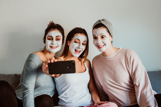 Group of friends applying a revitalizing white mask on their faces. Beauty treatment and skin care. Seated on the sofa taking pictures with a smpartphone together. Lifestyle.