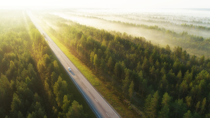 Aerial view of a highway with cars covered in fog. Early misty morning. Beautiful forest and sun rays.  Spotted from above with a drone. Finland, Europe. - 323993293