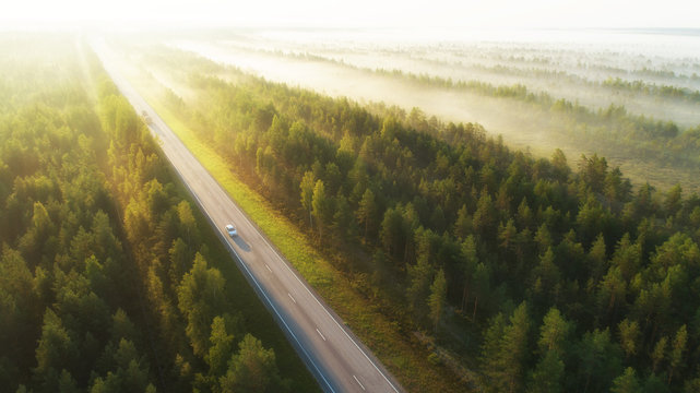 Aerial view of a highway with cars covered in fog. Early misty morning. Beautiful forest and sun rays.  Spotted from above with a drone. Finland, Europe.