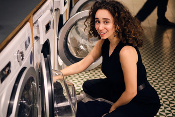 Fototapeta na wymiar Pretty brunette girl happily looking in camera taking out clothes from washing machine in modern self-service laundry