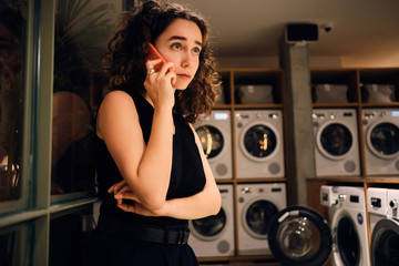 Beautiful girl talking on smartphone waiting washing in modern self-service laundry at night