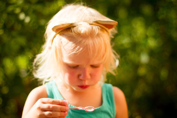 Pretty blond toddler girl wearing cat's headband making bubbles on a sunny summer day