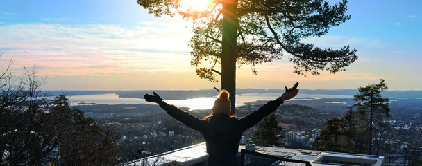 Happy feeling with a view of the City of Oslo, Norway in front. Oslo fjord in the background. Women standing with her hands in the air. Sunbeams glittering through a tree. - obrazy, fototapety, plakaty