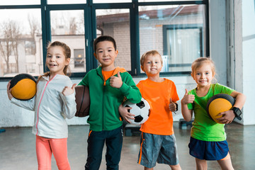 Front view of happy multicultural children holding balls in gym