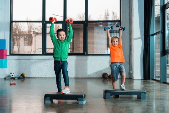 Front view of multiethnic children raising hands with dumbbells while doing Step Aerobics in gym