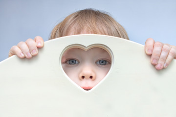 Baby boy toddler face, eyes and nose peep out of heart-shaped hole in center. A child looks into camera through a heart hole. His hands hold onto the fence with hole. Love and health concept.