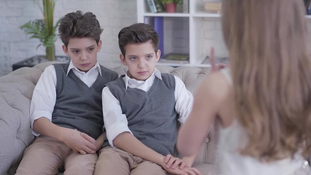 Unrecognizable little Caucasian brunette girl shaking finger as scolding twin brothers sitting on couch. Elegant boys shaking heads and looking at sister. Family, unity, lifestyle.