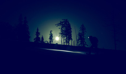 Winter mountain tourism. Night climbing of a tourist on snowshoes with a lamp on the head and a backpack. Moonlight illuminates the road. Light noise and dark night.