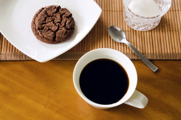 Morning black coffee in ceramic white cup, chocolate cookie on white plate, teaspoon, pen, bamboo placemat and sugar-bowl on table, messy arrangement, indoors, top view