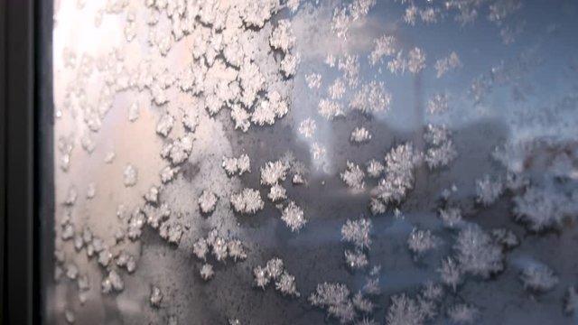 Hoarfrost ice crystals on window glass sparkling at sunrise. Beautiful icy flowers on frosty surface glittering in golden rising sun. Looking at sunset or dawn through frozen window against blue sky