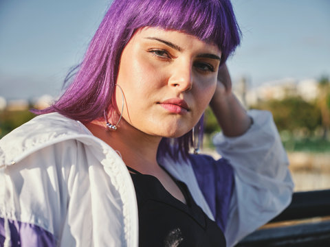 Fashion stylish woman with purple hairstyle touching face and leaning on metal fence in city center and confidently looking at camera in bright day