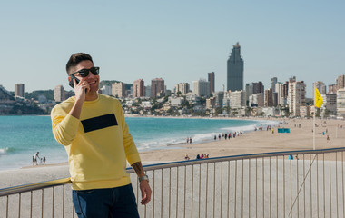 Satisfied young man in casual clothing and trendy sunglasses talking on mobile phone on waterfront with modern cityscape and beach on background