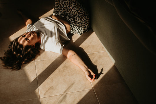 From above of tranquil woman lying on floor with spreader hands closed eyes and cigarette under rays of sun falling through window at home