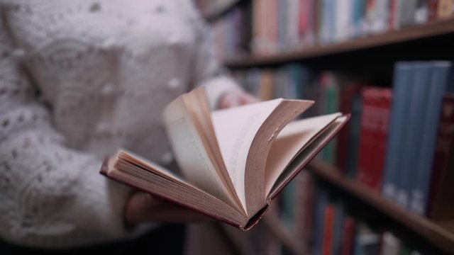  Unrecognizable woman beautifully turns over the pages of old paper book in big library on shelves background. Beautiful scene. Education, science, university concept.
