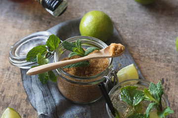 Overhead brown sugar in a jar near fresh limes and peppermint leaves placed on napkin on a wooden table